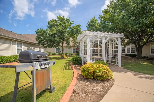 Outdoor area of Oxford Springs Tulsa Memory Care featuring a barbecue grill on a grassy lawn, a white wooden pergola structure, trees, bushes, and a paved walkway under a partly cloudy blue sky.
