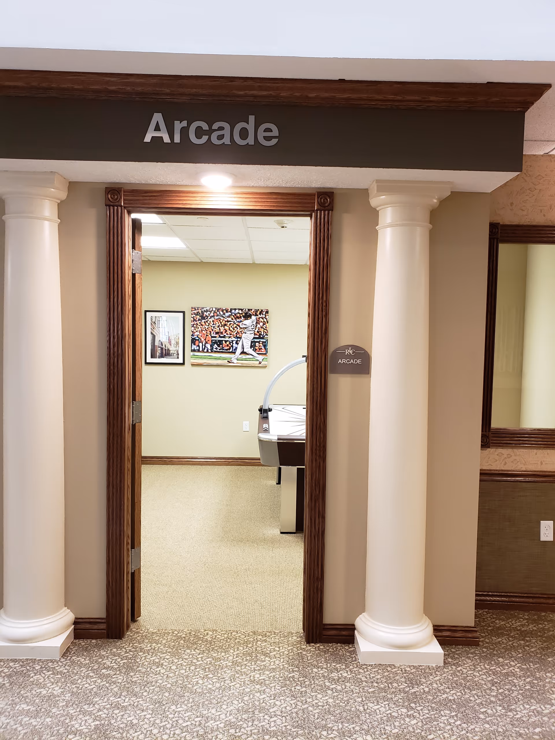 Entrance to a room labeled 'Arcade' with two white columns on either side of the doorway. Inside the room, there is an air hockey table and two framed pictures on the wall, one showing a baseball player in action.