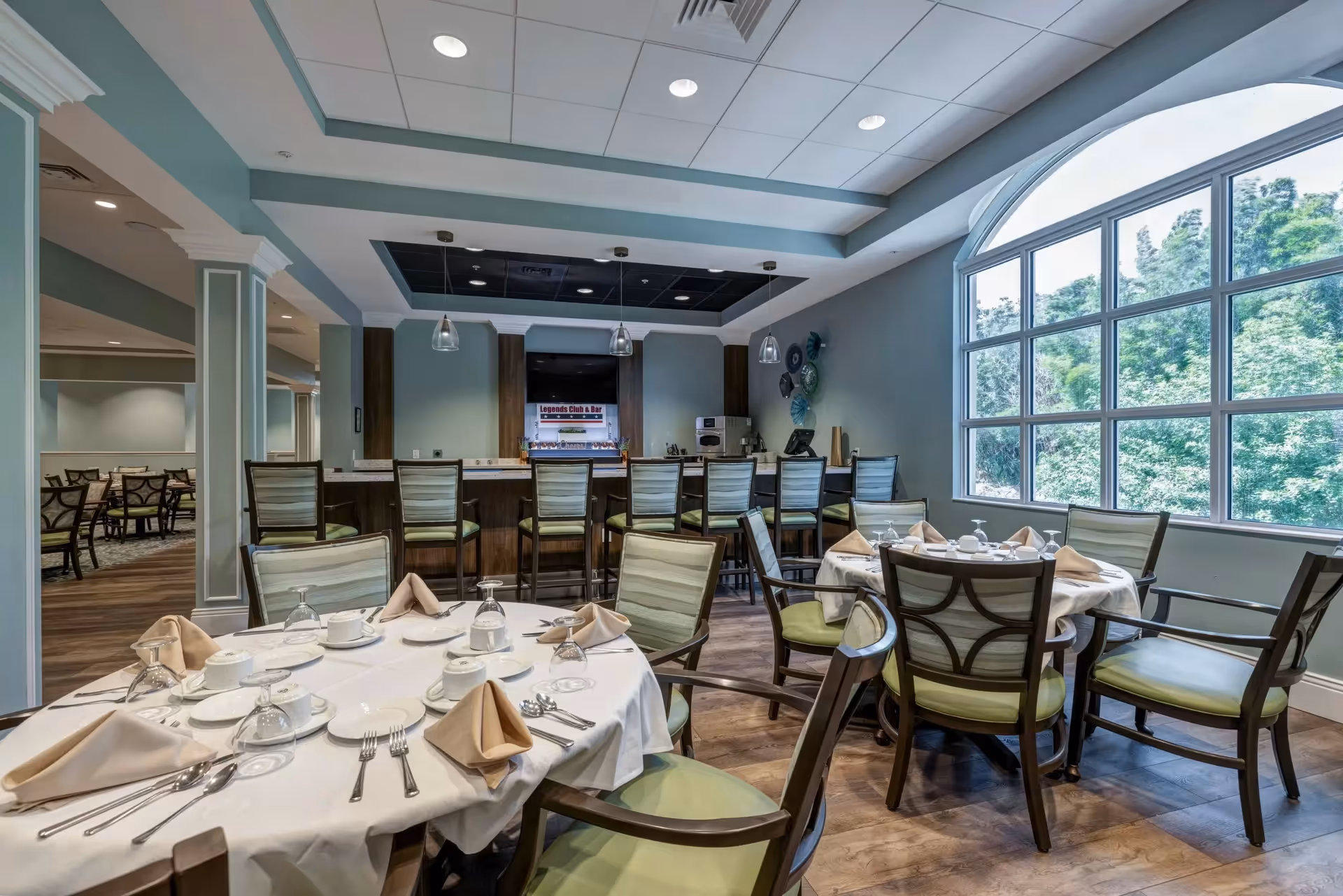 A dining room in a senior living facility with round tables set with white tablecloths, beige napkins, plates, cups, and silverware. There are green cushioned chairs around the tables. In the background, there is a bar area with high chairs and pendant lights hanging from the ceiling. Large windows on the right side let in natural light and show greenery outside.