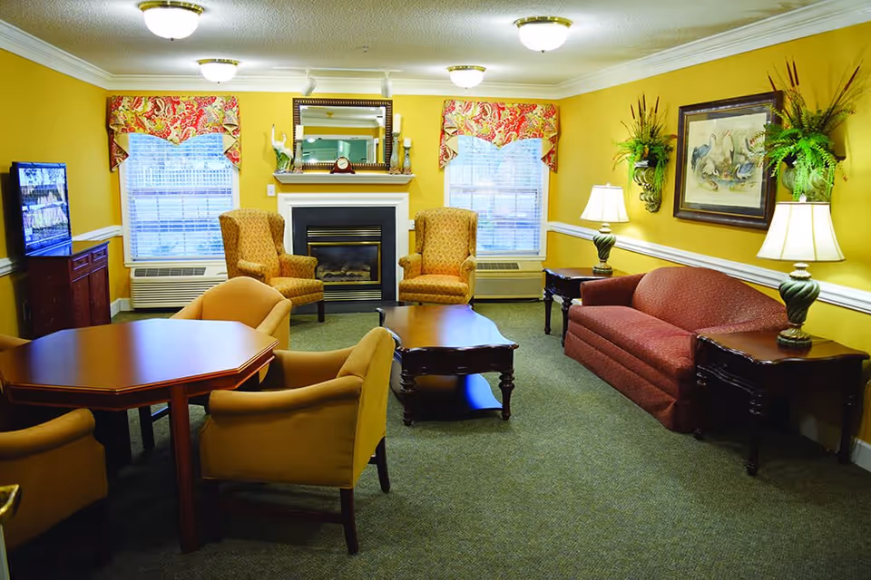 A cozy living room with yellow walls and green carpet featuring a red sofa, two patterned armchairs, a wooden coffee table, and a wooden dining table with four chairs. The room has two windows with floral valances, a fireplace with a mirror above it, two table lamps on side tables, and decorative plants and artwork on the walls.