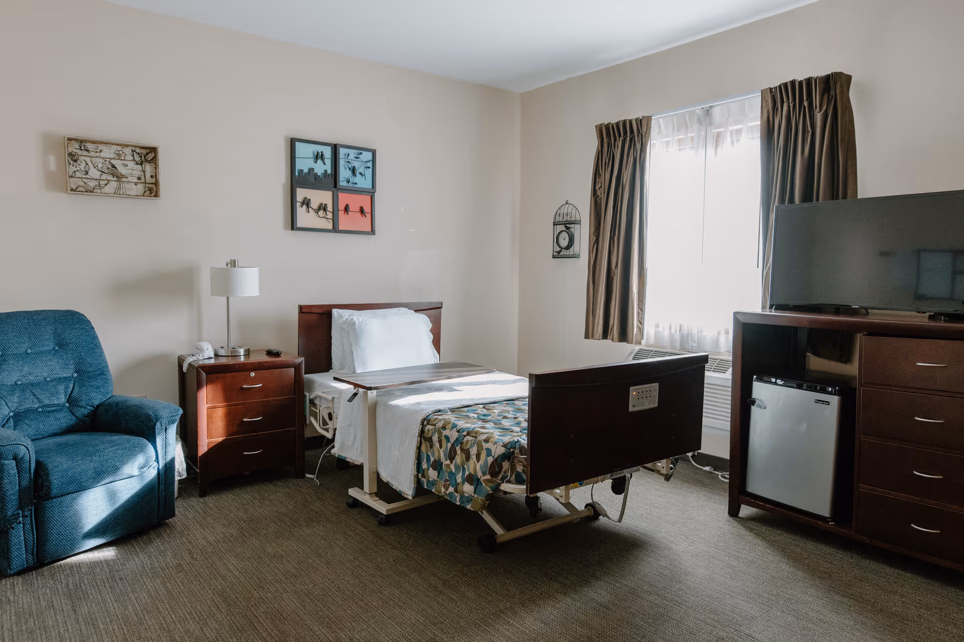 A well-lit bedroom in a senior living facility featuring a hospital-style bed with a patterned bedspread, a blue upholstered armchair, a wooden nightstand with a lamp and telephone, a window with brown curtains, and a wooden dresser with a flat-screen TV and mini refrigerator.