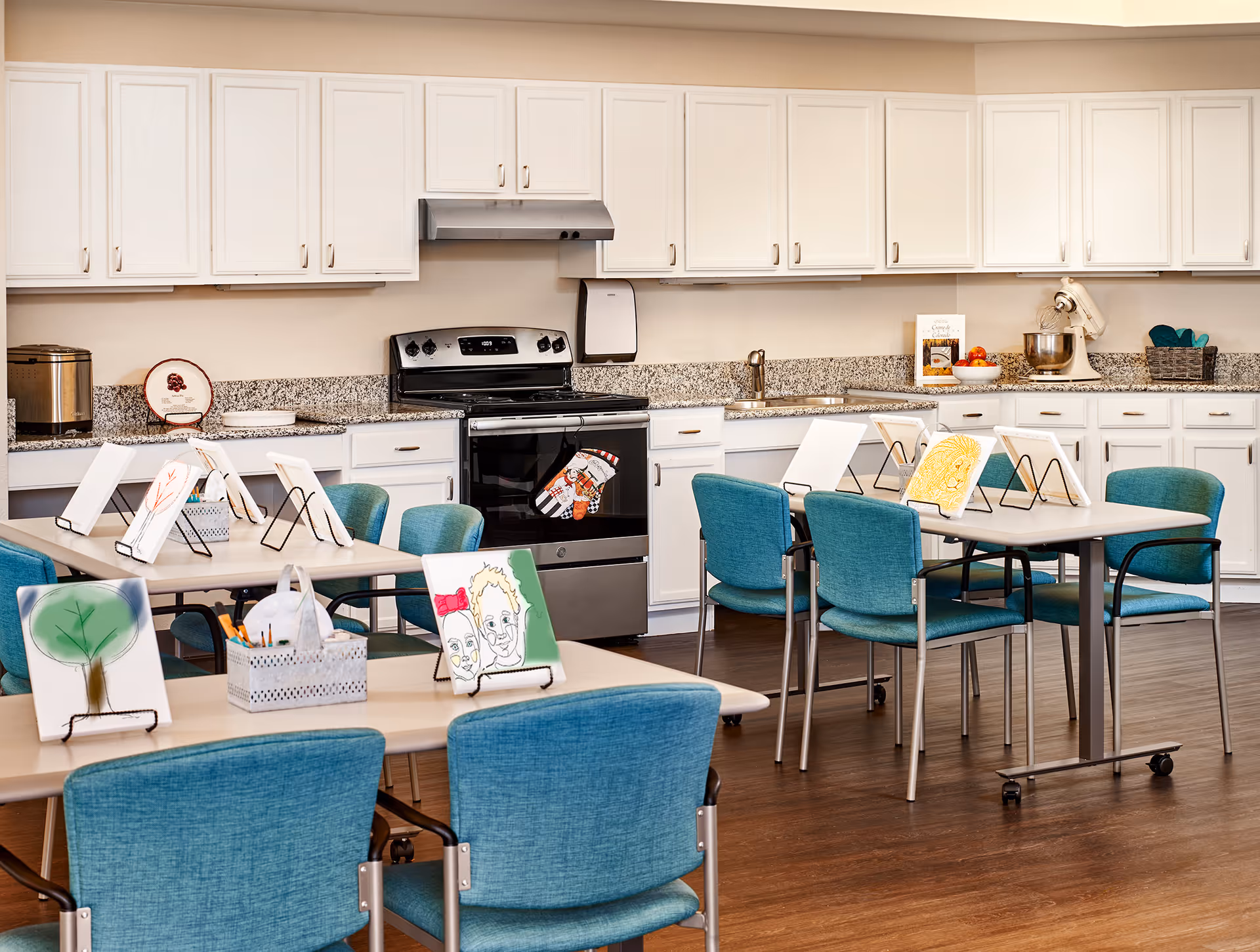 A bright and clean kitchen and activity room with white cabinets, granite countertops, and a stainless steel stove. Several tables with teal chairs are arranged in the room, each table displaying small canvases with colorful drawings and paintings. Art supplies are also visible on the tables.