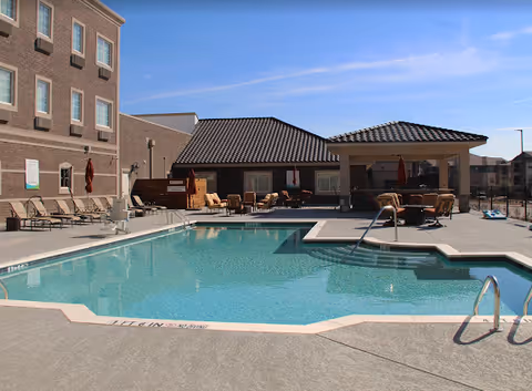 Outdoor swimming pool area at a senior living facility with lounge chairs, umbrellas, and a covered seating area. The pool is surrounded by a concrete deck and adjacent buildings with windows are visible in the background under a clear blue sky.