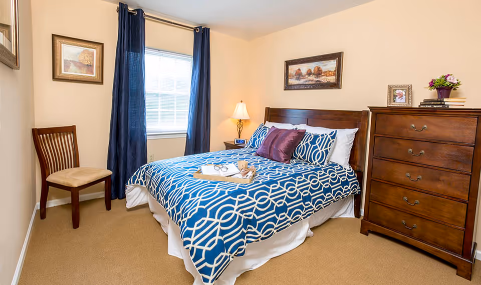 Well-lit bedroom with a double bed in blue patterned bedding, a wooden headboard and dresser, a chair, lamp, and a window with dark curtains.