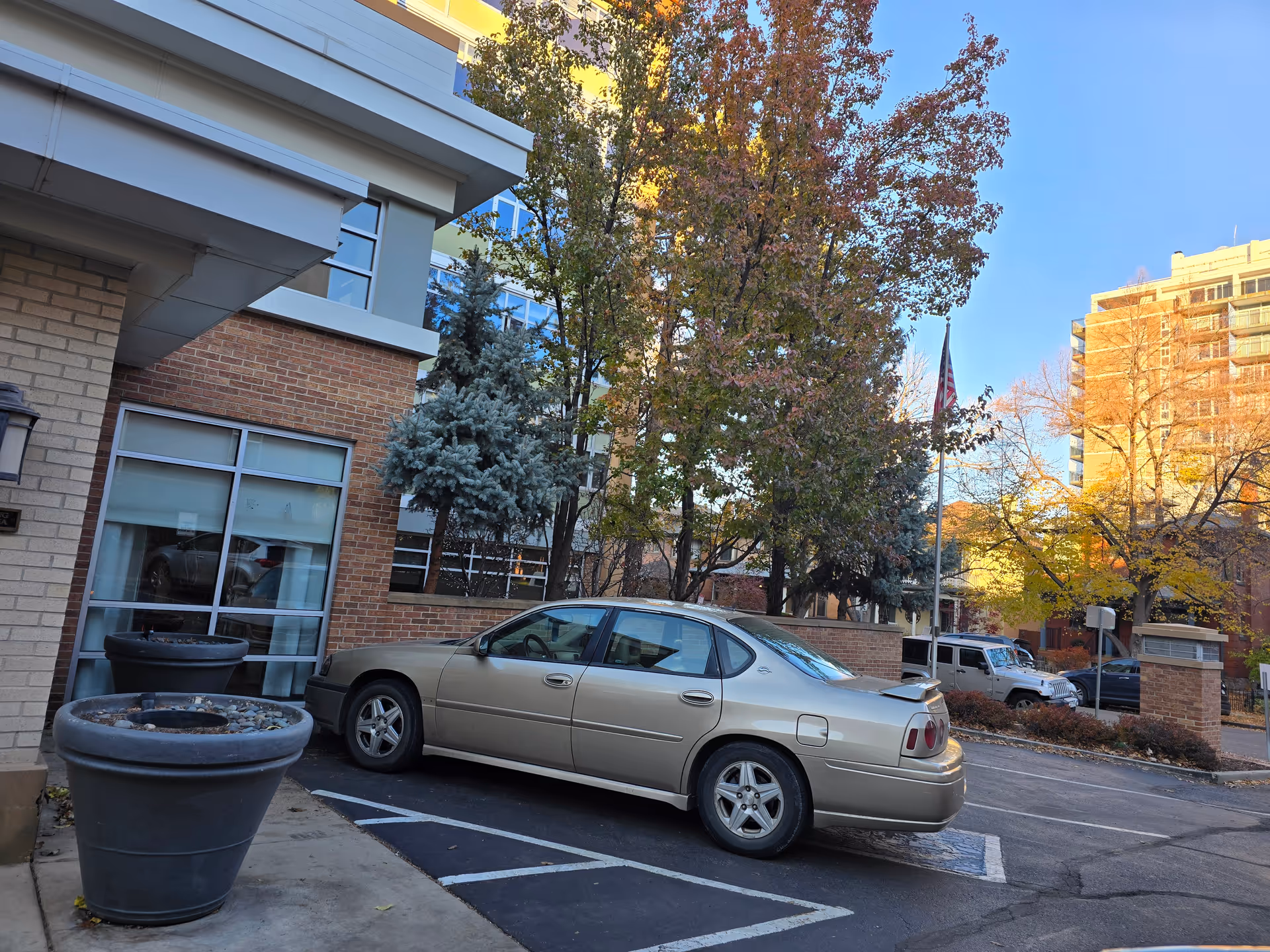 A beige sedan car parked in a parking lot next to a building with brick and beige walls. There are large empty planters on the sidewalk near the car. Trees with autumn foliage and a flagpole with an American flag are visible in the background, along with other buildings under a clear blue sky.