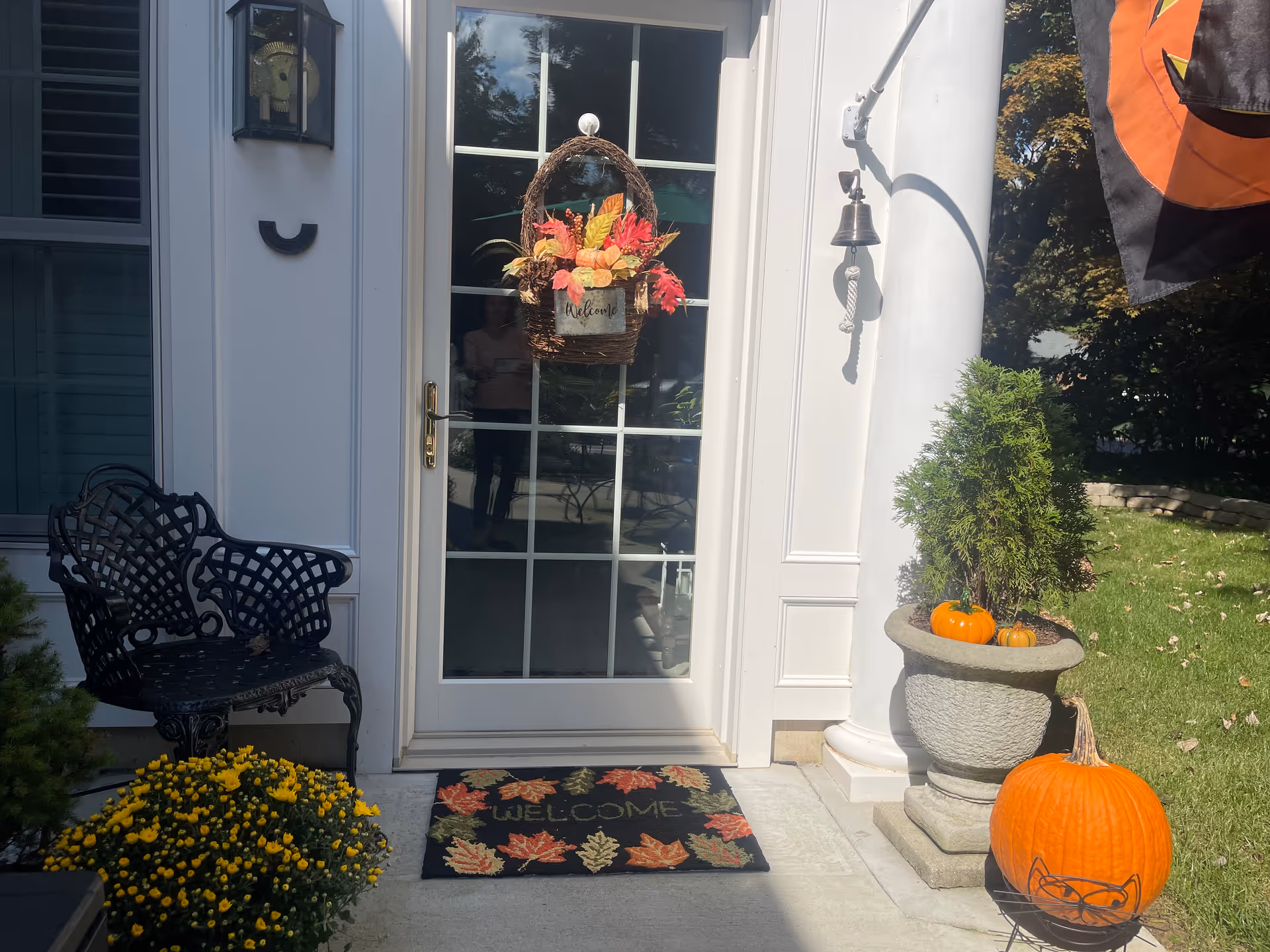 Front entrance of a house with a glass door decorated with a fall-themed wreath and a welcome mat featuring autumn leaves. There is a black metal chair to the left, a potted plant with small pumpkins on the right, and a large pumpkin on the ground. A small bell hangs on a white column next to the door, and part of a Halloween flag is visible on the right side.