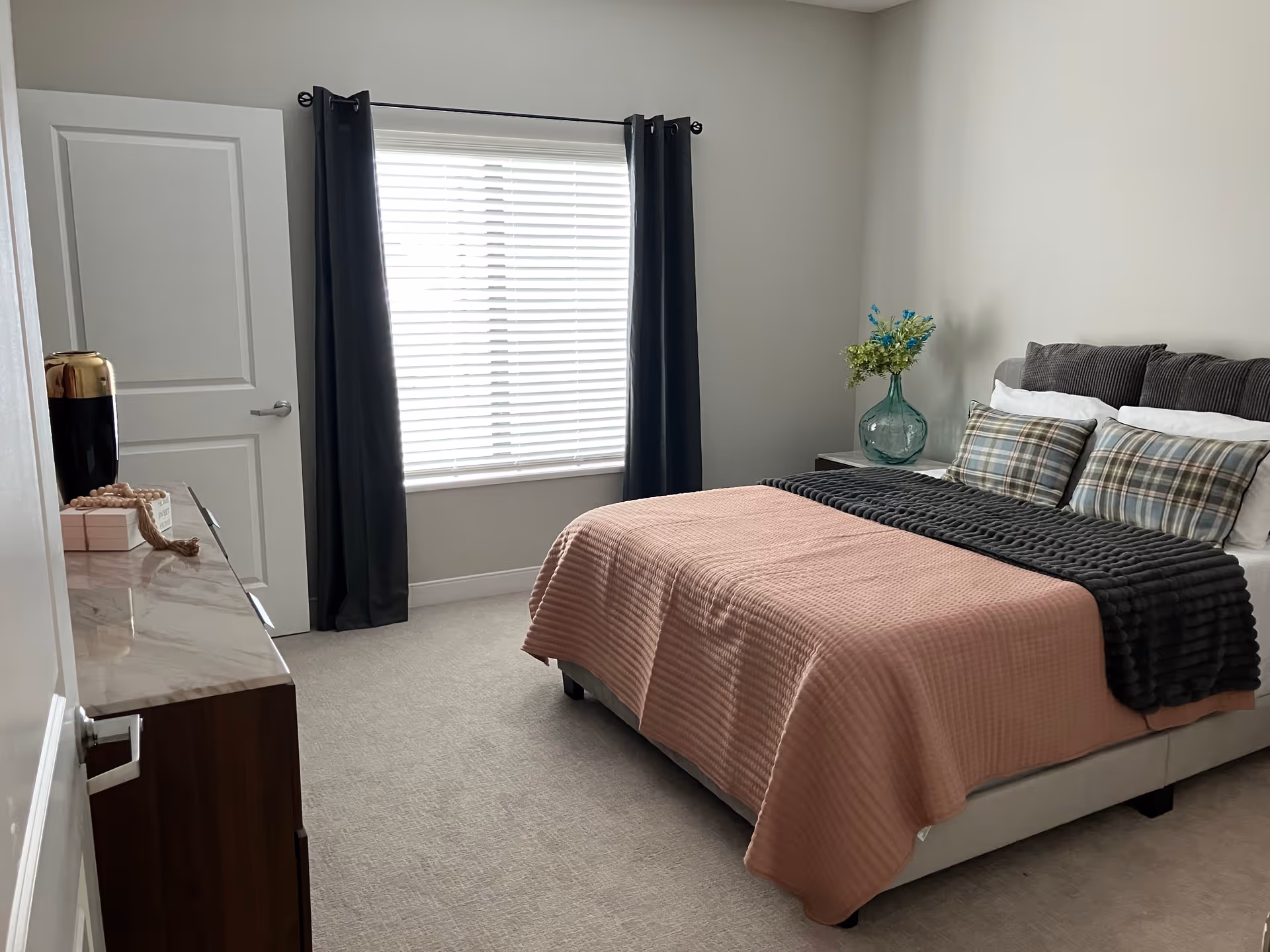 Sunlit bedroom with a made bed featuring a pink quilt and gray throw, a window with blinds and dark curtains, a dresser, and a vase of flowers on a nightstand.