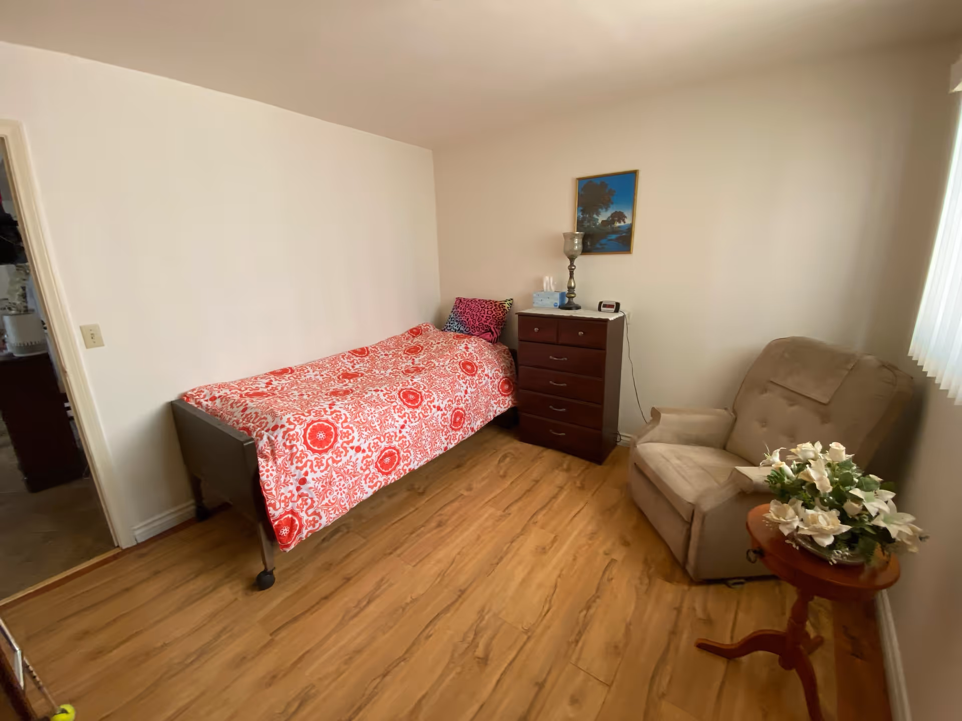 Small bedroom with a single bed covered in red patterned bedding, a wooden dresser, a beige recliner, and a small side table with flowers.