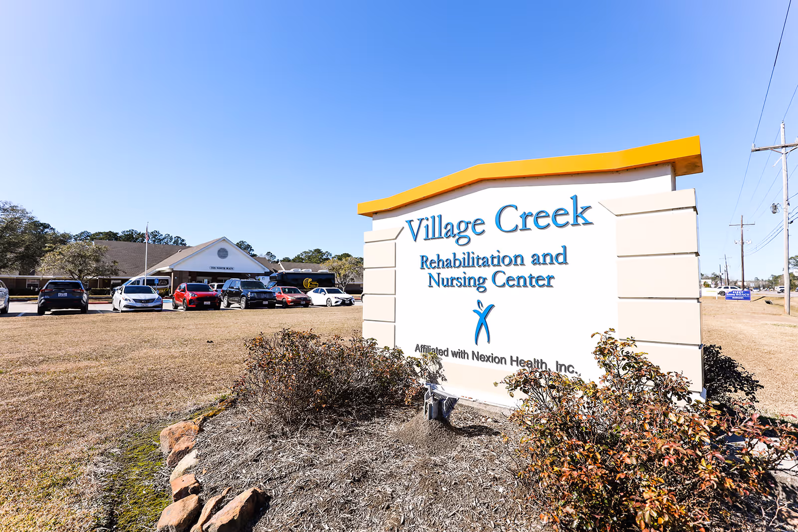 Outdoor view of Village Creek Rehabilitation and Nursing Center sign with the building and parked cars in the background under a clear blue sky.