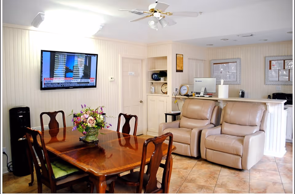 A cozy common area in an assisted living facility featuring a wooden dining table with six chairs and a floral centerpiece. On the wall, a flat-screen TV is mounted showing a news channel. Two beige recliner chairs are positioned near a reception desk with a computer and office supplies. The room has tiled flooring, light-colored walls, and a ceiling fan.