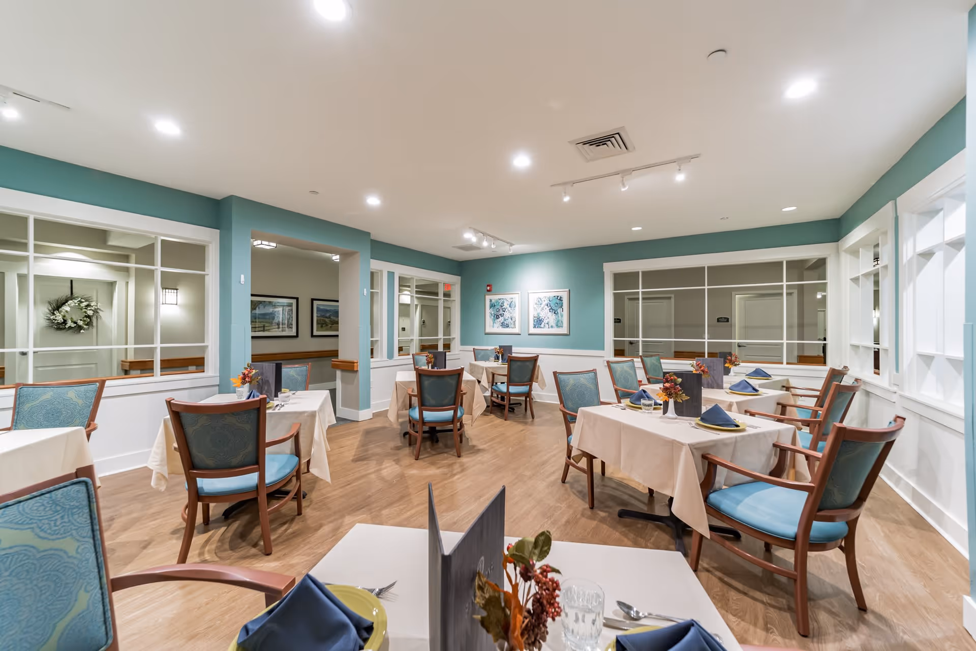A bright and clean dining room with several tables covered in white tablecloths, each set with blue napkins, plates, glasses, and silverware. The chairs have wooden frames with blue upholstered seats and backs. The walls are painted teal with white wainscoting, and there are large windows and framed artwork on the walls. The floor is wood, and the ceiling has recessed lighting and track lights.