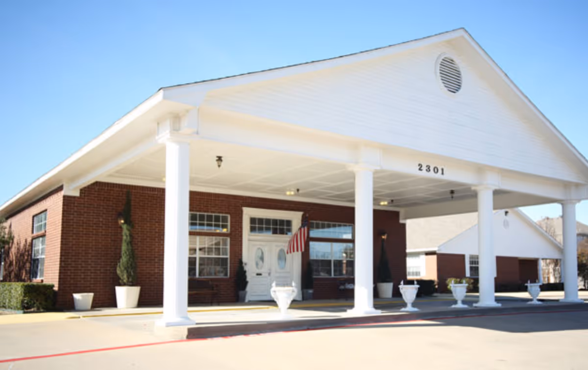 Front exterior view of a brick building with white columns supporting a large covered entrance. The building has the number 2301 displayed above the entrance, an American flag near the door, and several potted plants along the front. The sky is clear and blue.