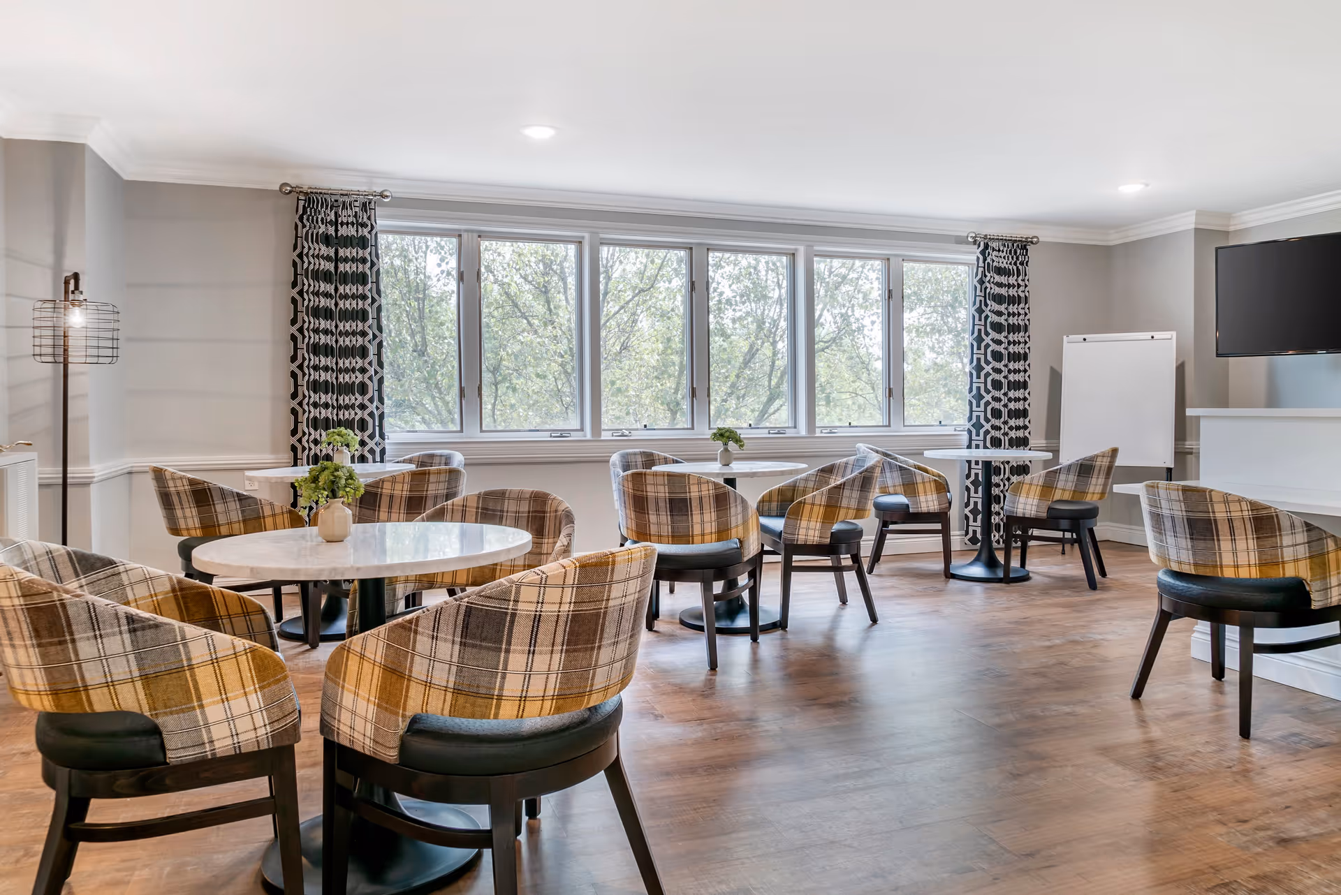 A bright and spacious common area with several round tables surrounded by plaid upholstered chairs. Large windows with patterned curtains let in natural light and offer a view of trees outside. The room has wooden flooring, a flat-screen TV mounted on the wall, a whiteboard, and a modern floor lamp in the corner.