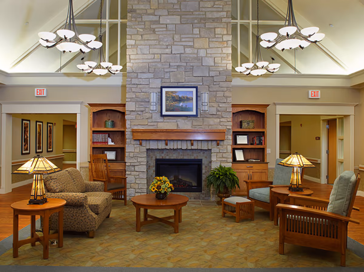Well-lit common living room with chairs and tables arranged around a central stone fireplace and chandeliers overhead.