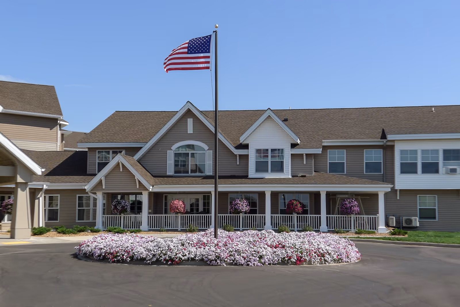 Front exterior view of The Deerfield facility with a circular flower bed filled with white and pink flowers surrounding a flagpole flying the American flag. The building has beige siding, white trim, multiple windows, and a covered porch with hanging flower baskets.