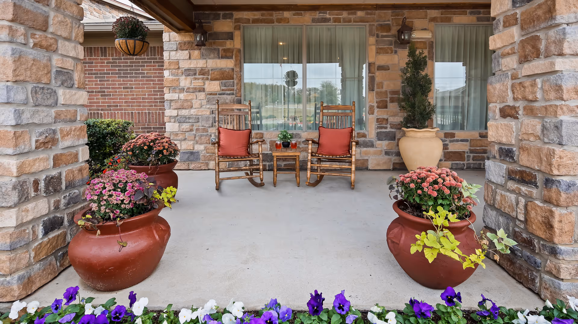 Covered outdoor patio area with two wooden rocking chairs with red cushions and a small wooden table between them. The patio is surrounded by large stone pillars and walls, with several large red pots containing pink flowers and green plants. In the foreground, there are purple and white flowers along the edge of the patio.