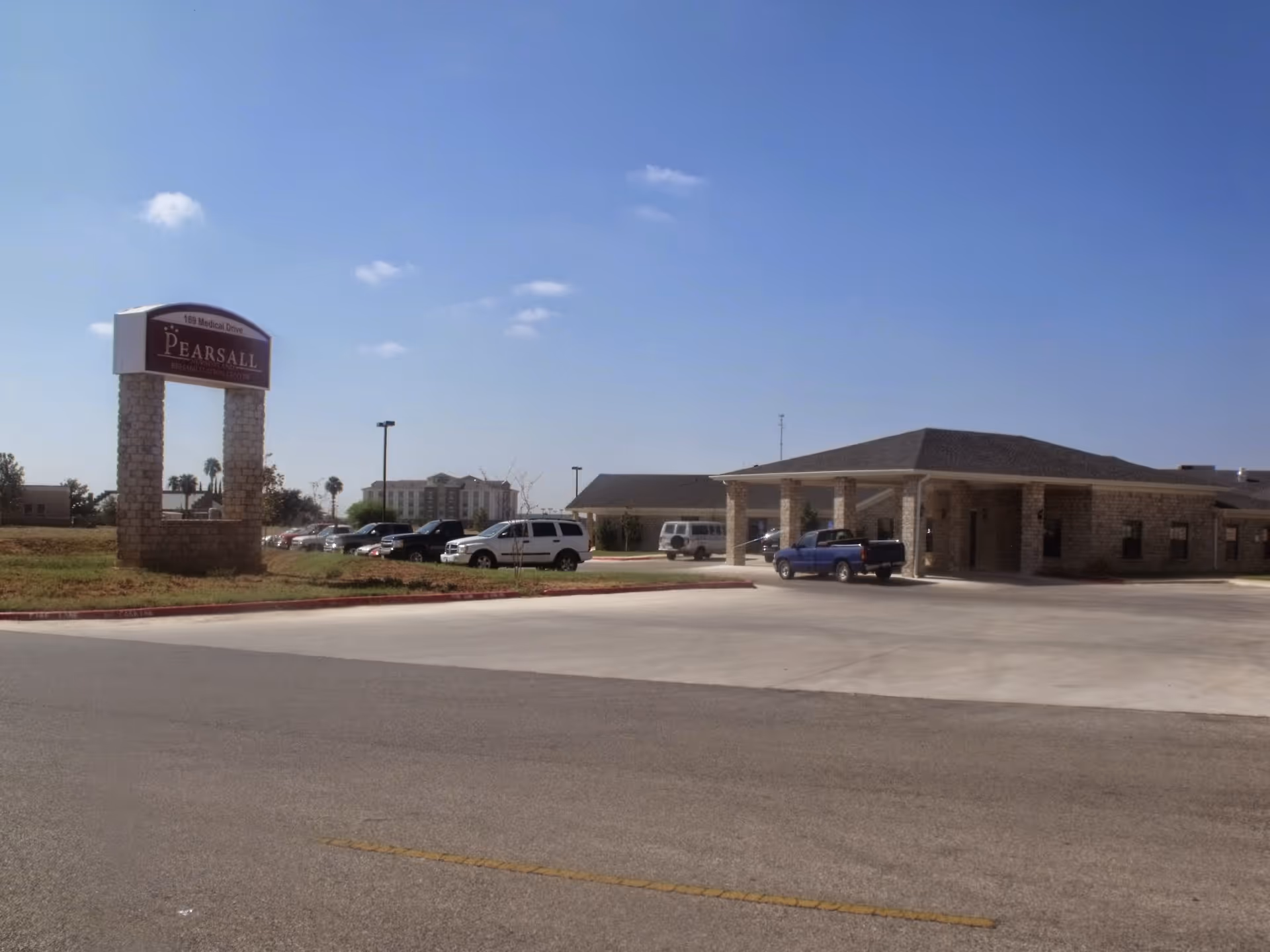 Exterior view of Pearsall Nursing and Rehabilitation Center showing a stone sign with the facility name and address, a parking lot with several vehicles, and a single-story building with a covered entrance under a clear blue sky.