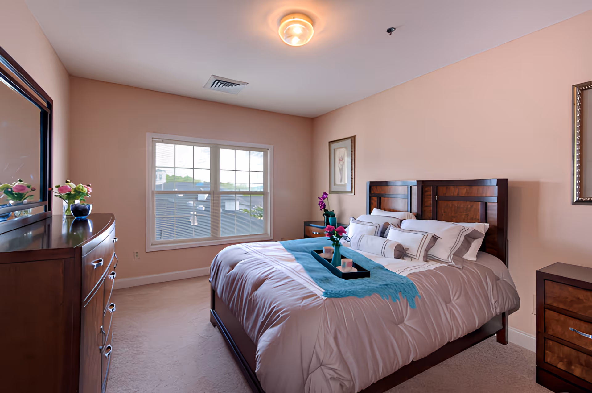 A neatly made bed with a wooden headboard in a bedroom with peach-colored walls. The bed has white and gray bedding with multiple pillows and a teal throw blanket. There is a wooden dresser with a mirror on the left side and a nightstand with a vase of flowers on the right side. A window with white trim lets in natural light, and framed artwork hangs on the walls.