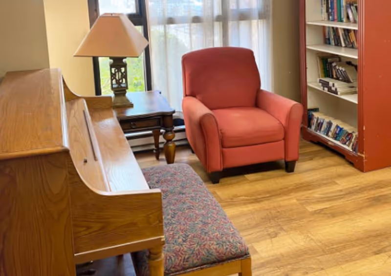 A cozy interior room featuring a wooden piano with a patterned bench, a wooden side table with a lamp, a red upholstered armchair, and a bookshelf filled with books. The room has wooden flooring and a window with sheer curtains allowing natural light to enter.