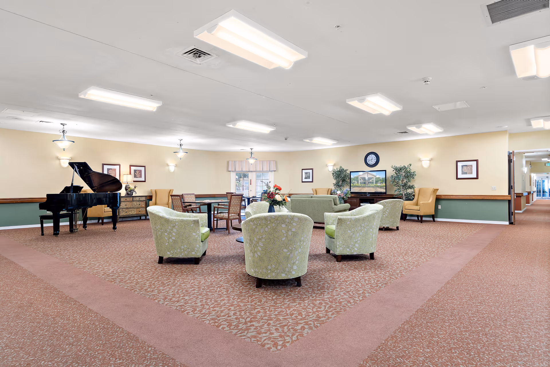 A spacious senior living facility common area with patterned carpet and soft lighting. The room features a grand piano on the left, several green upholstered armchairs arranged around a small table with a flower arrangement, and a few yellow wingback chairs along the walls. There is a TV mounted on the far wall with plants on either side, framed artwork, and a clock above the TV. A hallway extends to the right side of the room.