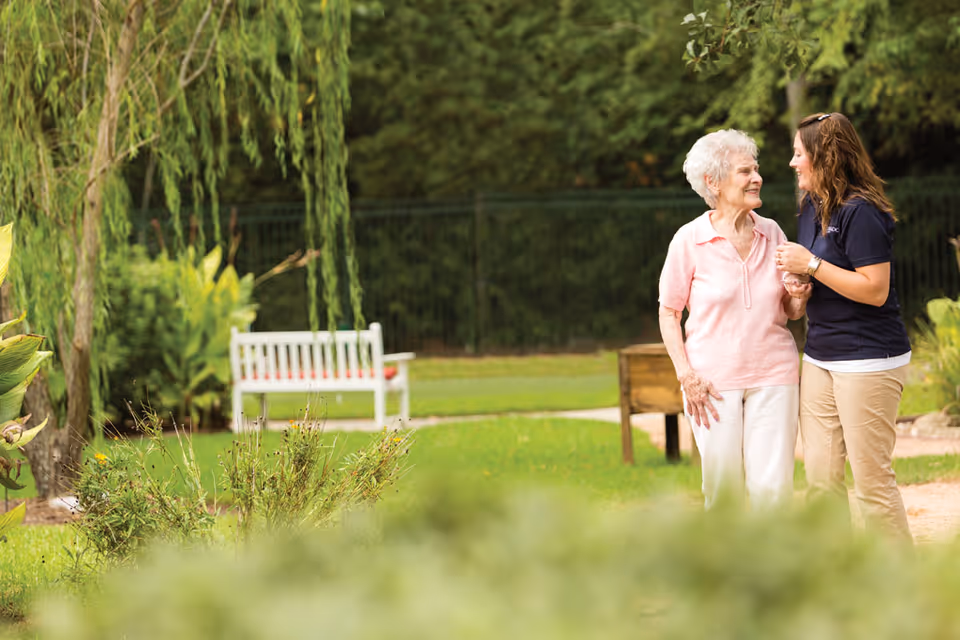 An elderly woman and a younger woman are walking together outdoors in a garden area. The elderly woman is wearing a light pink shirt and white pants, while the younger woman is dressed in a navy blue shirt and beige pants. They are smiling and holding hands, surrounded by greenery, plants, and a white bench in the background.