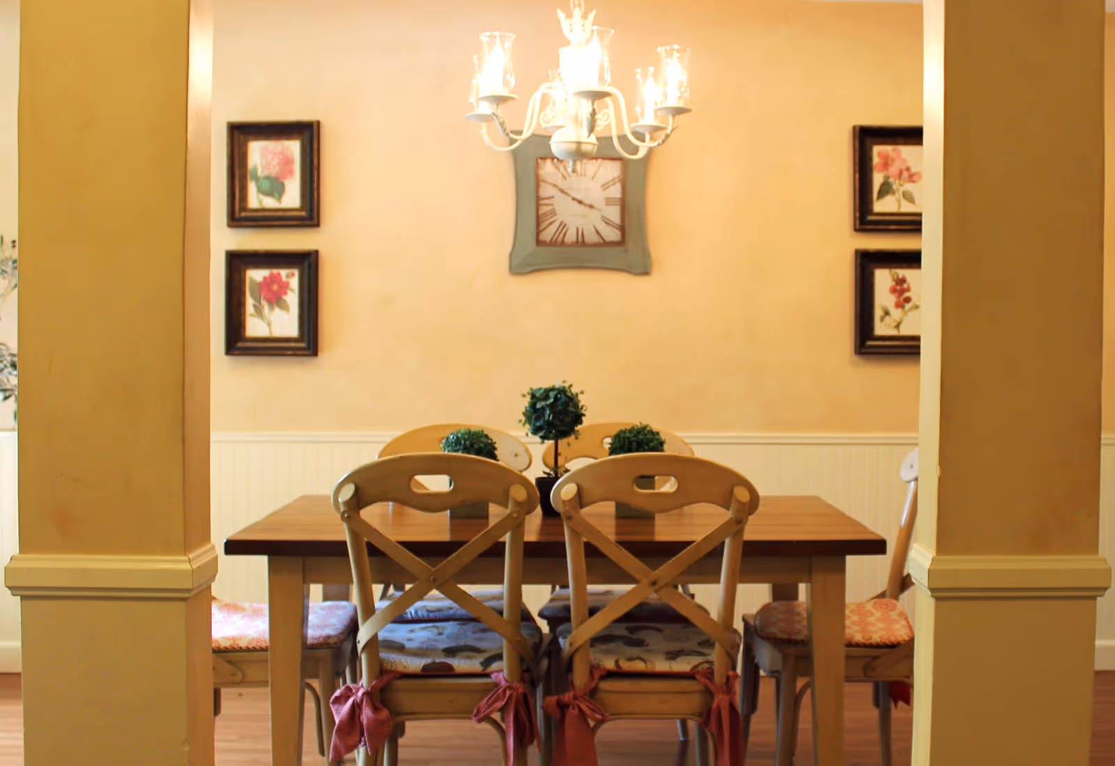 A cozy dining room with a wooden table and six chairs, each with patterned cushions tied to the seats. The walls are painted a warm beige color and decorated with framed floral artwork and a wall clock. A chandelier with multiple lights hangs above the table, and small green potted plants are placed on the table as centerpiece decorations.