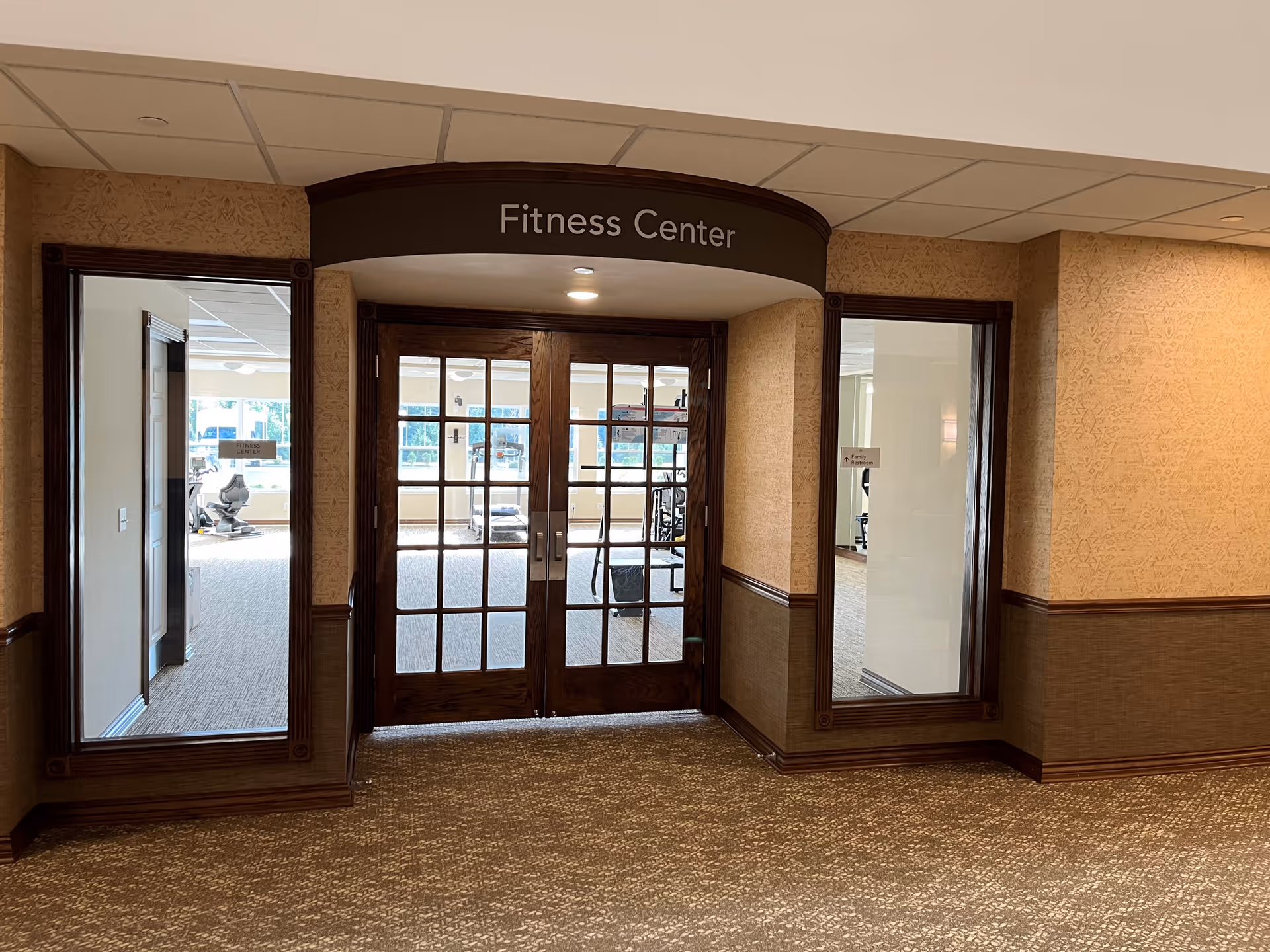 Entrance to a fitness center with double wooden framed glass doors and windows on either side, showing exercise equipment inside. The area outside the fitness center has patterned carpet and beige walls with wood trim.