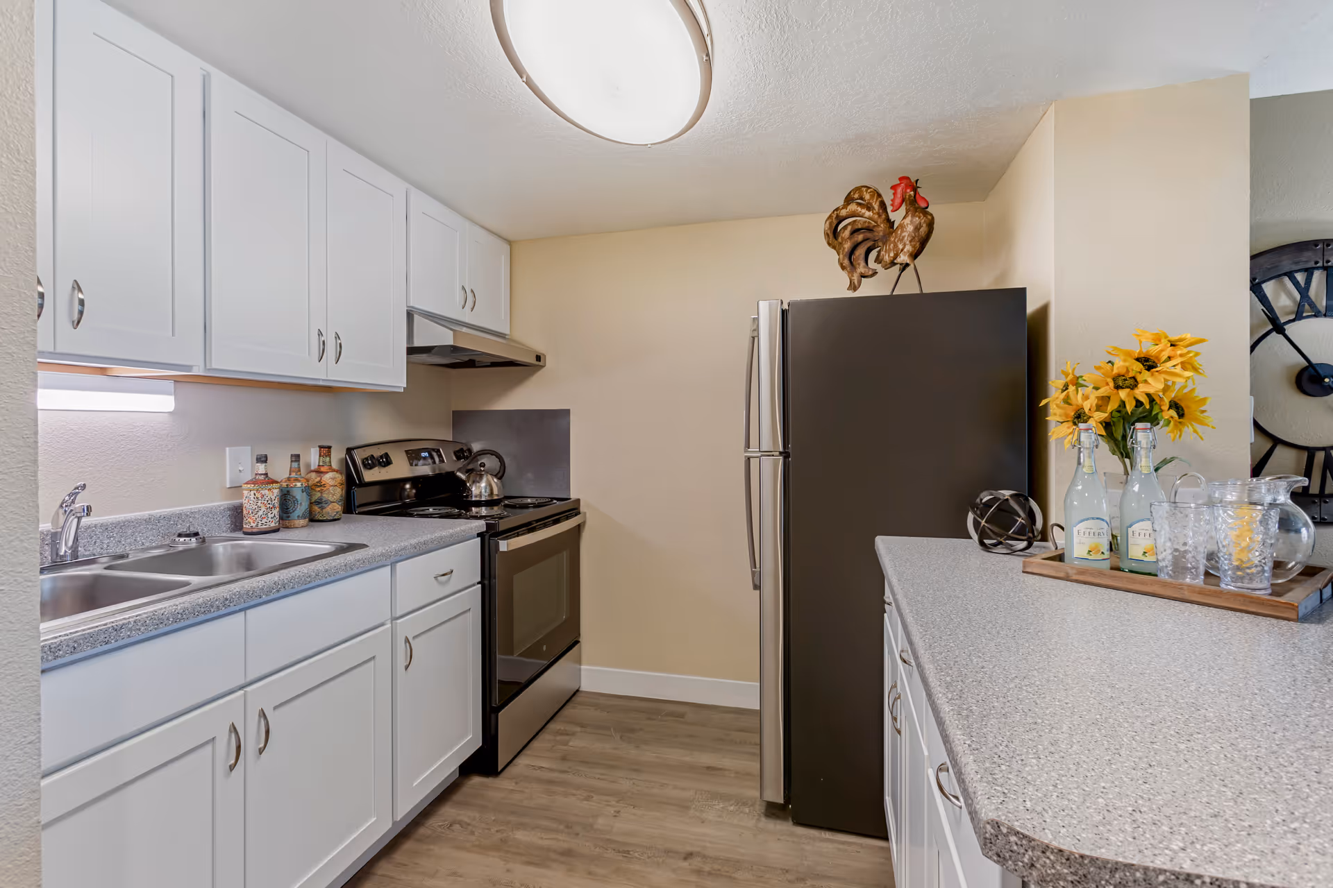A modern kitchen with white cabinets, a stainless steel stove and refrigerator, a double sink, and a countertop decorated with bottles, a rooster figurine, and a tray holding flowers and glassware.