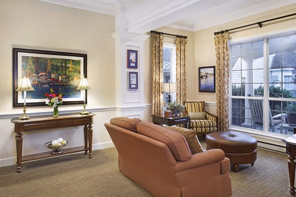 A cozy living room with a brown sofa, a striped armchair, a round leather ottoman, and a wooden side table with a lamp and plant. There is a large window with patterned curtains showing an outdoor porch with rocking chairs. A wooden console table with two lamps and a vase of flowers is against the wall, beneath a framed painting of a lakeside village.