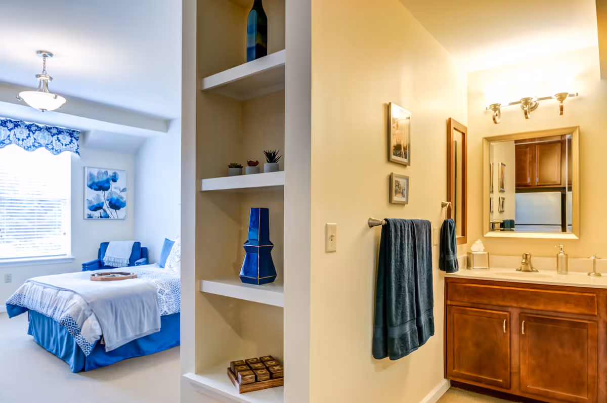 A bright and cozy bedroom with a blue and white color scheme is visible through an open doorway. The bedroom features a bed with blue bedding, a blue armchair, a window with a blue floral valance, and a wall painting of blue flowers. Adjacent to the bedroom is a bathroom vanity area with a wooden cabinet, a sink, a mirror, and a towel rack holding blue towels. Between the bedroom and bathroom is a built-in shelving unit displaying decorative items including small plants and blue vases.