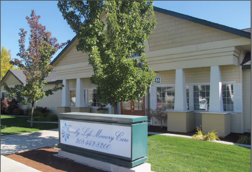 Exterior view of a single-story building with beige siding and white trim, surrounded by green grass and trees. In front of the building is a sign that reads 'Family Life Memory Care 208-442-8200'.