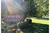 Brick entrance sign reading 'TRINITY VILLAGE' surrounded by landscaping and trees with sunlight streaming through.