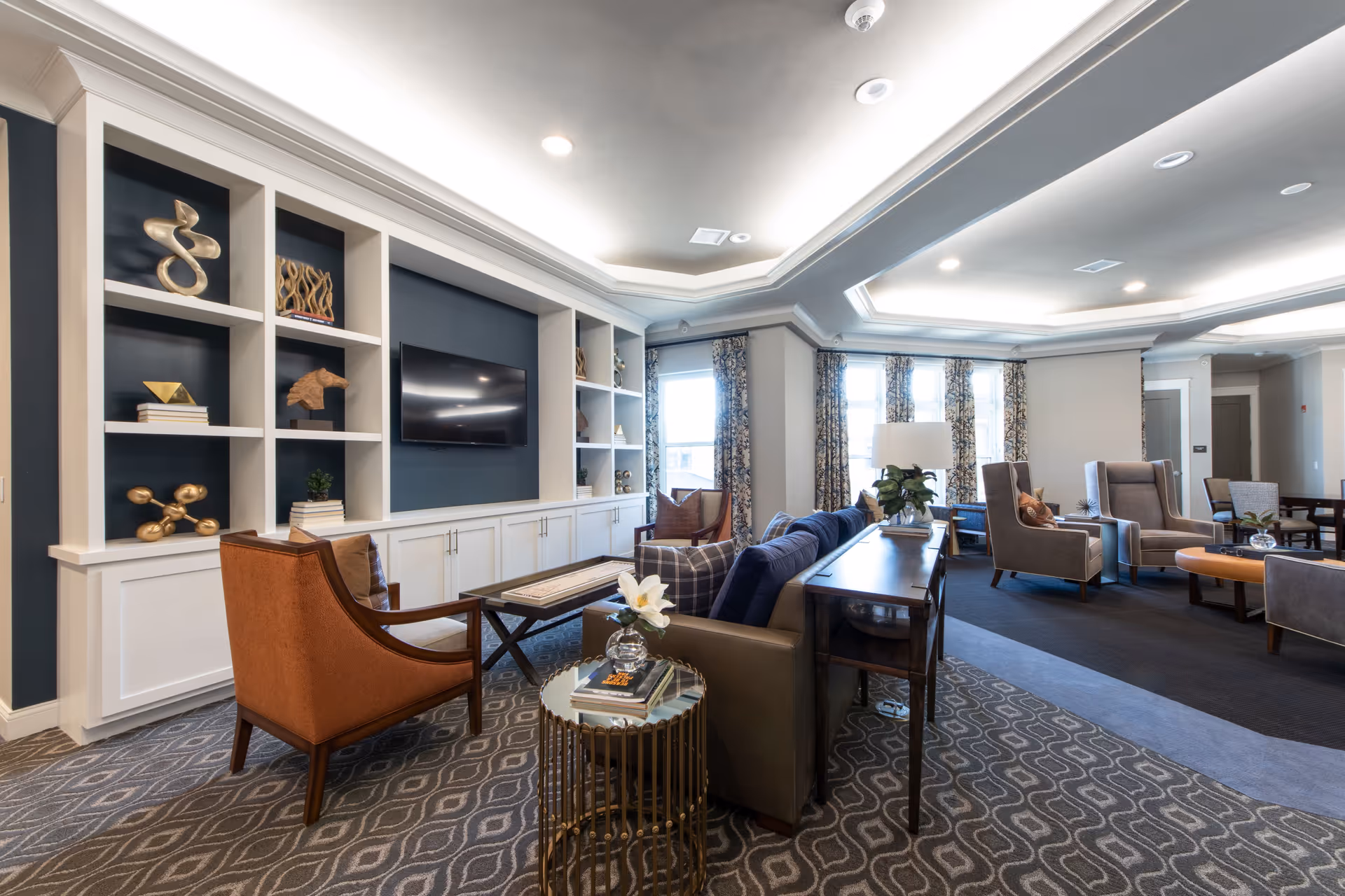 Bright upscale common living room with built-in shelving and TV, sofas and armchairs arranged around coffee tables.