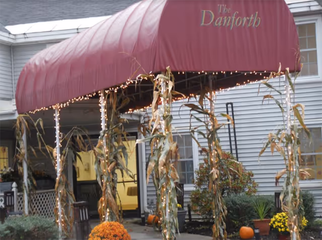 Front entrance of The Danforth adult care center with a burgundy canopy, string lights, corn stalk decorations, pumpkins and potted mums.