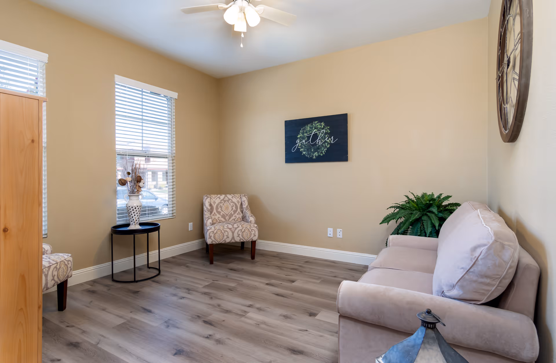 A cozy living room with beige walls and wood flooring featuring a beige sofa with a large cushion, a patterned armchair, a small round black side table with a decorative vase, a green potted plant, a large wall clock, and a wall art piece with the word 'gather' and a wreath design. Two windows with white blinds allow natural light into the room.