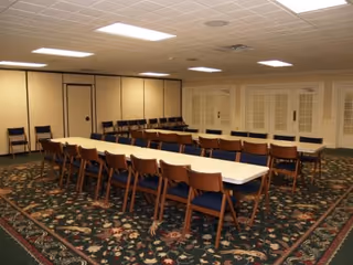 A large meeting or dining room with two long white tables arranged end to end, surrounded by wooden chairs with blue cushions. The room has a patterned carpet, beige walls, and a drop ceiling with fluorescent lights. Folding chairs are stacked against the far wall.