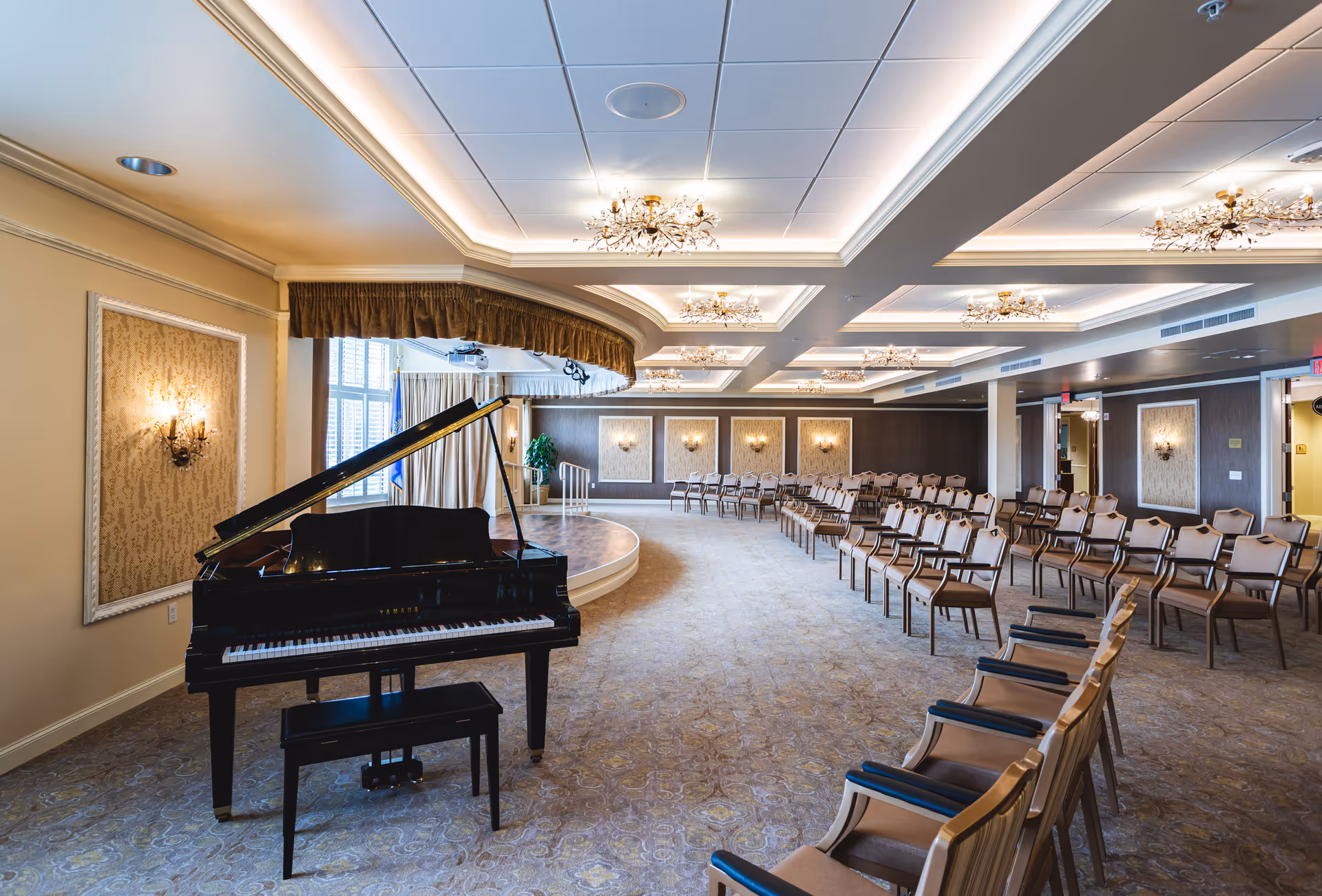 A spacious, elegant room with a grand piano in the foreground and rows of chairs arranged facing a small stage with a podium. The room features ornate wall sconces, decorative ceiling lights, and large windows with curtains, creating a formal and inviting atmosphere.
