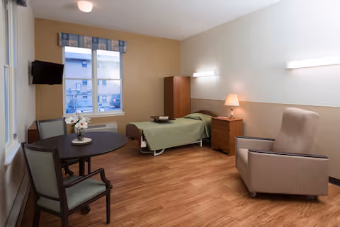 A simple, clean bedroom in a senior living facility with a single bed covered in a green blanket, a wooden nightstand with a lamp, a wooden cabinet, a round table with two chairs, a beige armchair, and a window showing an outside view of a building and parked cars.