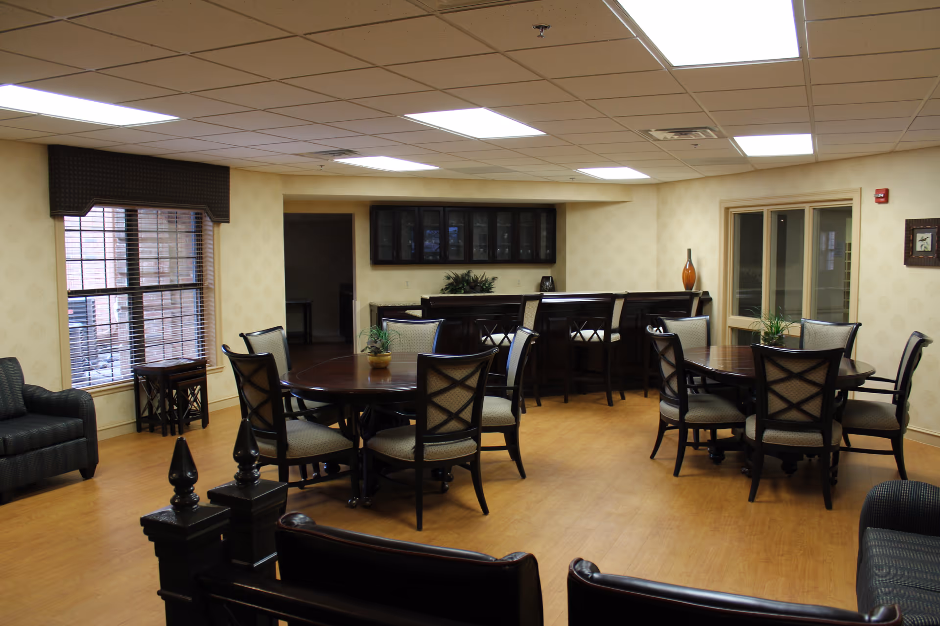 A senior community common area with two round wooden tables surrounded by cushioned chairs. There is a bar counter with high chairs in the background, a window with blinds on the left, and a few couches and small tables around the room. The floor is wooden, and the ceiling has recessed lighting panels.