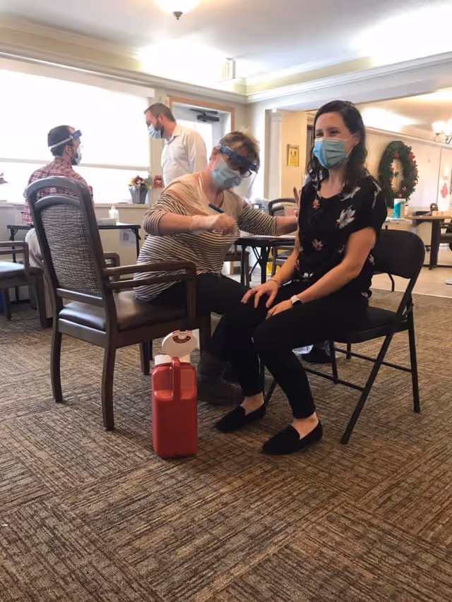 A healthcare worker wearing a face mask and face shield is administering a vaccine or injection to a seated woman who is also wearing a face mask. Two other masked individuals are visible in the background sitting and standing in a well-lit room with carpeted flooring and chairs.