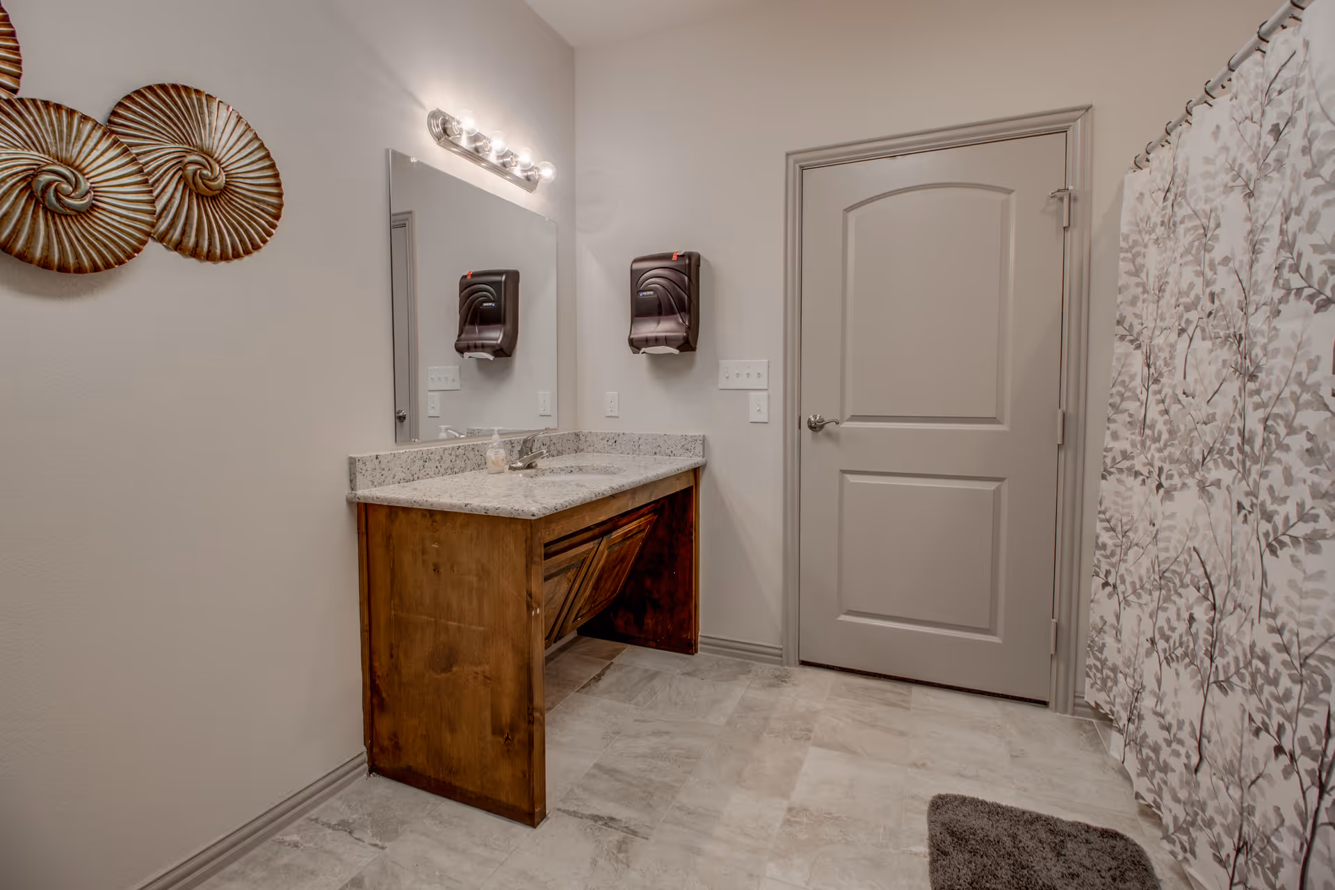 Light-colored bathroom with a granite countertop vanity, wall mirror, closed door, and patterned shower curtain.