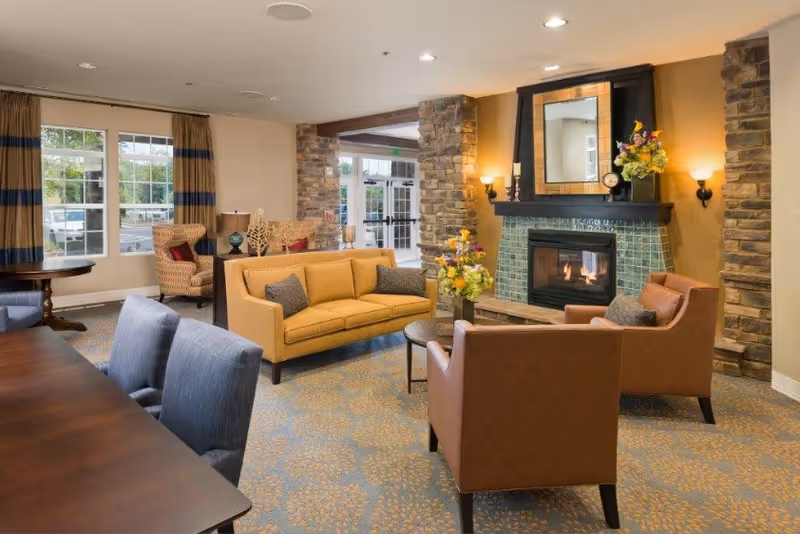 A cozy living room area with a yellow sofa, two brown armchairs, a round coffee table with a flower arrangement, and a fireplace with a tiled surround and a mirror above it. The room has stone pillars, large windows with curtains, and a carpeted floor with a floral pattern.