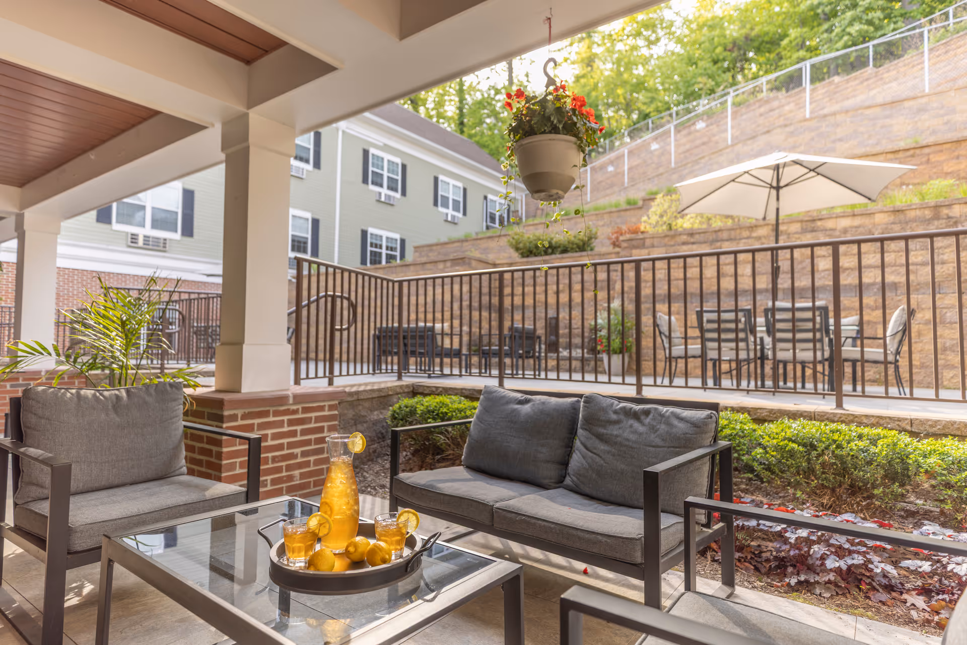 Outdoor patio area with cushioned seating including a sofa and chairs around a glass coffee table holding a tray with a pitcher of iced tea, glasses, and lemons. The patio is covered with a ceiling and supported by white columns. In the background, there is a raised area with additional seating and a large white umbrella, along with a retaining wall and greenery.
