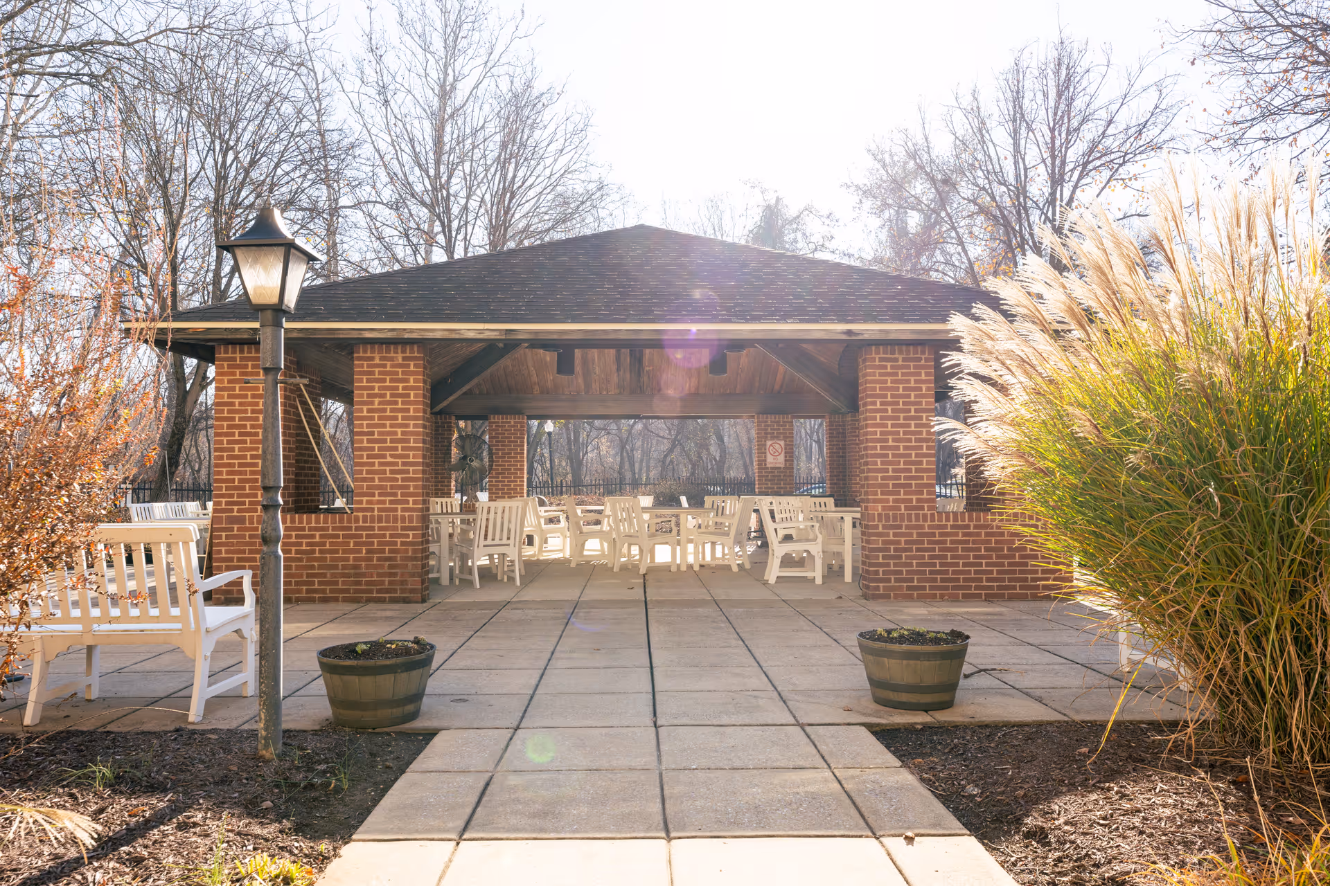 Outdoor covered seating area with a brick structure and a dark shingled roof. White benches and chairs are arranged under the shelter. There are two large planters on either side of the paved walkway leading to the seating area. Surrounding the area are leafless trees and ornamental grasses, indicating a late autumn or winter season.