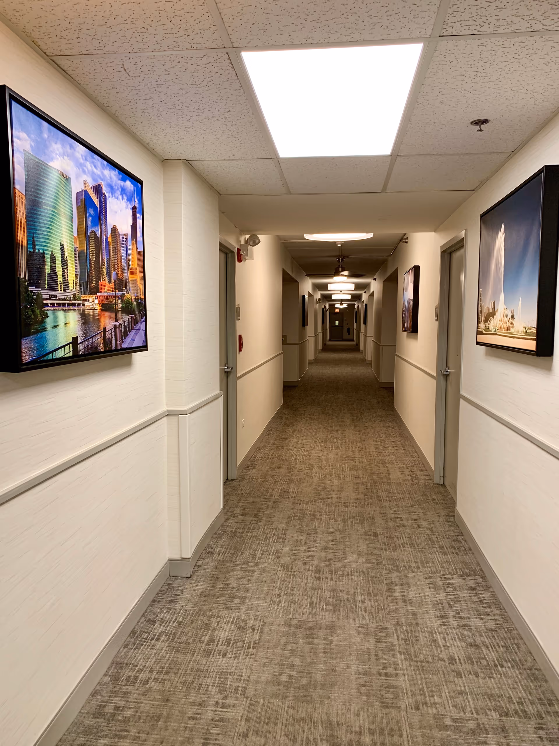 A long, well-lit hallway in a supportive living facility with beige walls and carpeted floor. The hallway has several closed doors on both sides and framed pictures of cityscapes hanging on the walls. Ceiling lights illuminate the corridor evenly.