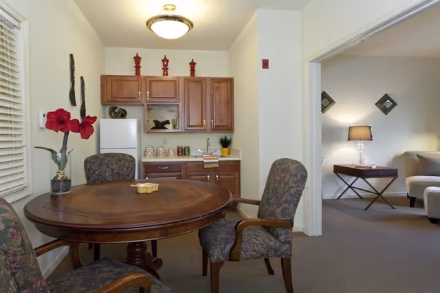 Small dining area with a round wooden table and upholstered chairs facing a kitchenette, opening into a cozy living room.