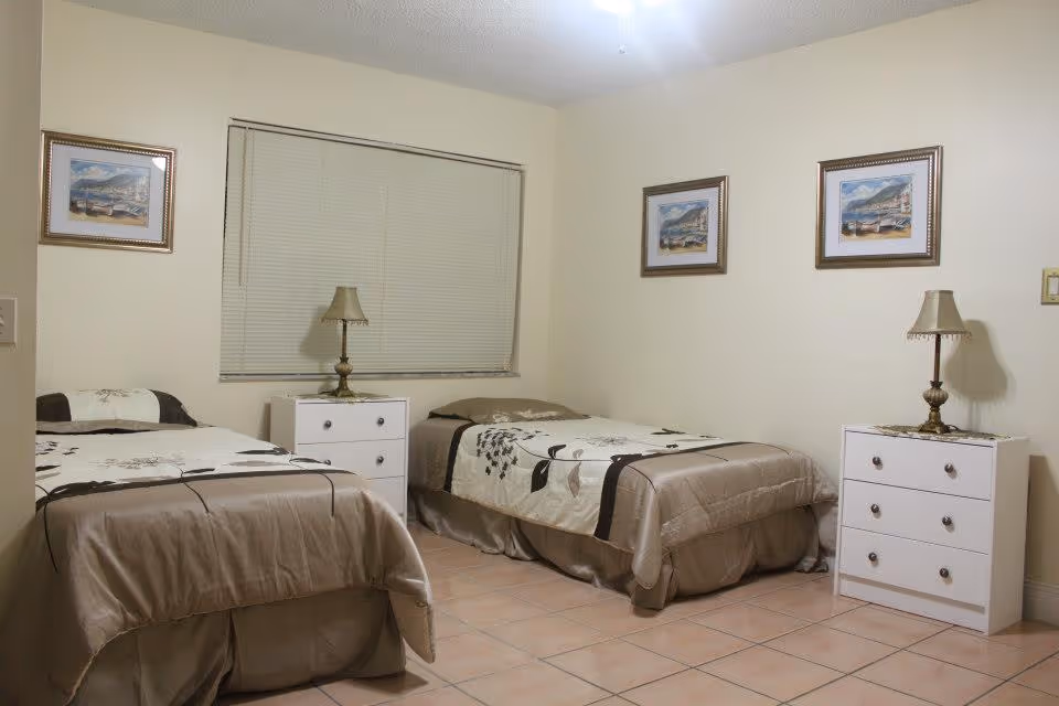 Bedroom with two twin beds, matching white nightstands and lamps, and framed pictures on beige walls.