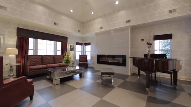 A spacious living room with patterned wallpaper, a large window with dark red curtains, a patterned sofa, two armchairs, a modern rectangular coffee table with a flower arrangement, a wall-mounted electric fireplace, and a grand piano with decorative items on top.