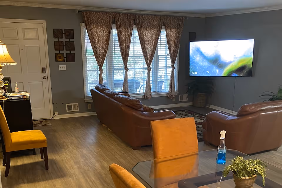 Cozy living room with leather couches, a wall-mounted TV, large window with patterned curtains, and a glass-top table with chairs.