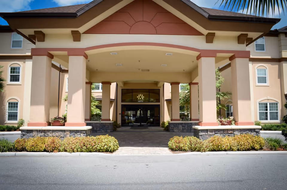Front entrance of a senior living facility with a covered driveway supported by large beige columns with stone bases, surrounded by landscaped bushes and plants. The building has multiple windows and a peaked roof with a clear blue sky above.